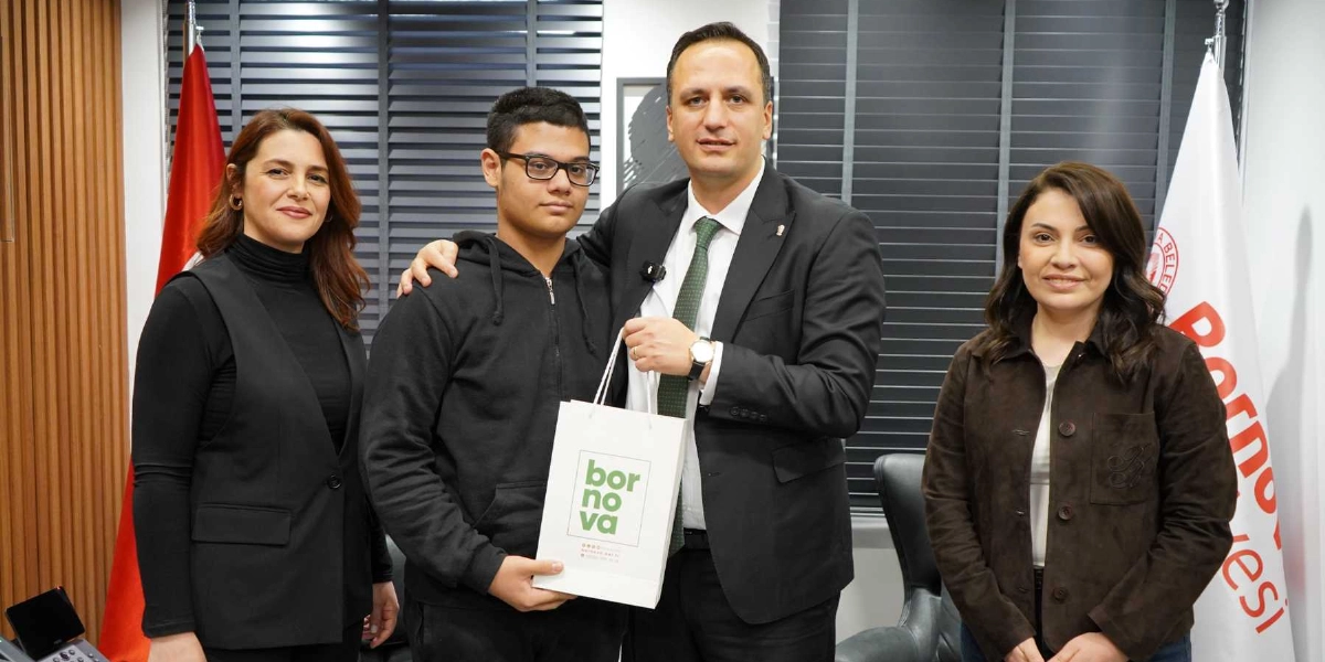 Bornova Belediye Başkanı, siyah kapüşonlu sweatshirt giyen gözlüklü bir genç öğrenciye üzerinde Bornova Belediyesi logosu bulunan beyaz bir hediye çantası veriyor. Yanlarında iki kadın yetkili bulunuyor; arka planda ise Türk bayrağı ve Bornova Belediyesi flaması yer alıyor.