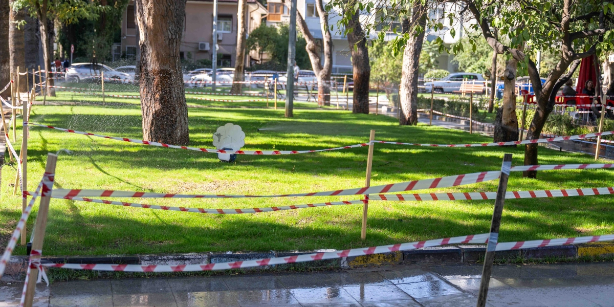 Büyükpark'ta, etrafı kırmızı beyaz ikaz şeritleriyle çevrilmiş yeşil çim alanın ortasında, otomatik sulama sistemiyle çimlere su püskürten bir fıskiye çalışıyor. Arka planda ağaçlar, parkın kenarında park halindeki araçlar ve binalar görülüyor.