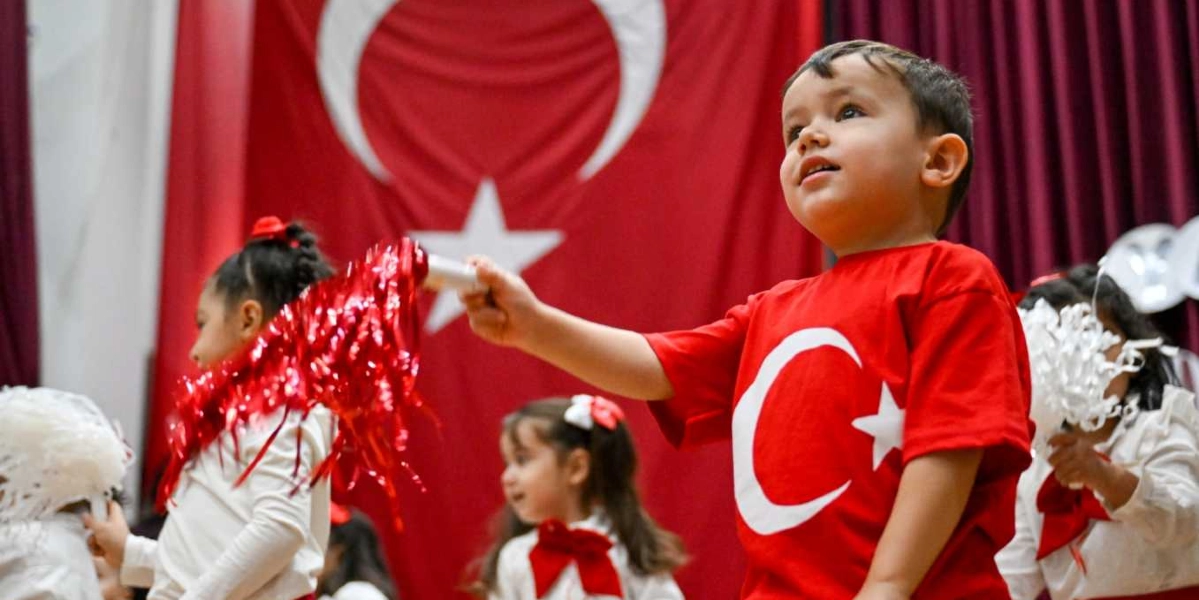 Arka planda büyük bir Türk bayrağının asılı olduğu sahnede, üzerinde Türk bayrağı baskılı kırmızı tişört giyen küçük bir erkek çocuk elinde kırmızı bir ponpon tutarak neşeyle ileriye bakıyor. Çocuğun çevresinde beyaz kıyafetler giymiş, ellerinde ponponlar bulunan diğer çocuklar Cumhuriyet Bayramı kutlaması için gösteri yapıyor.