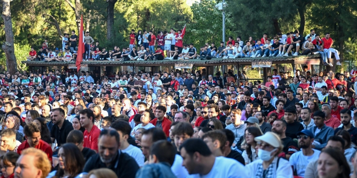 Ağaçlık bir alanda, bir etkinliği izlemek için toplanmış çok sayıda insan. Kalabalığın bir kısmı, arkadaki kapalı bir yapının çatısına oturmuş, bir kısmı ise ön tarafta ayakta veya oturarak sahneye doğru bakıyor. Ortamda yoğun bir kalabalık hakim ve insanlar dikkatle bir noktaya odaklanmış durumda.