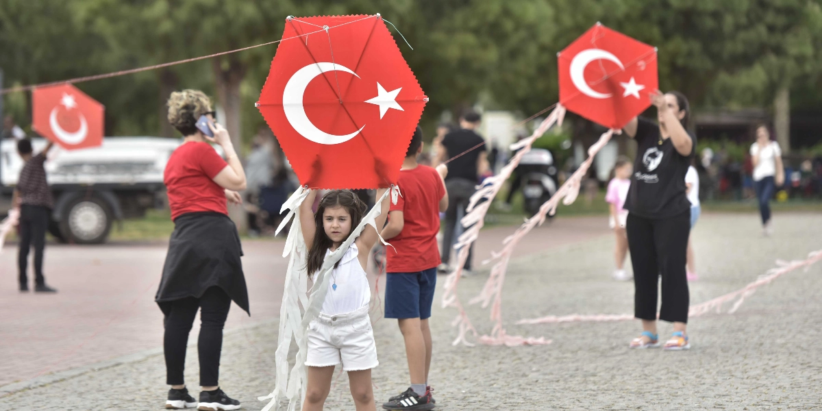 Açık havada, park benzeri bir alanda, üzerinde Türk bayrağı deseni bulunan altıgen uçurtmalar tutan çocuklar ve onlara eşlik eden yetişkinler. Ön planda, beyaz kıyafetli küçük bir kız çocuğu, üzerinde ay yıldız bulunan kırmızı uçurtmayı iki eliyle başının üzerinde tutuyor. Arka planda ise benzer uçurtmalar tutan başka çocuklar, telefonla konuşan bir kadın ve yürüyen insanlar görülüyor.