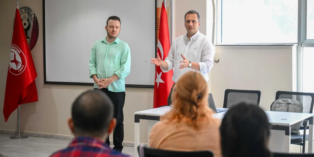 Aydınlık bir toplantı salonunda, beyaz bir gömlek giymiş bir konuşmacı ellerini kullanarak katılımcılara hitap ediyor. Yanında açık yeşil gömlekli bir kişi ayakta duruyor. Arka planda Bornova Belediyesi logosunun bulunduğu bir bayrak ve Türk bayrağı yer alıyor. Ön planda ise toplantıyı dinleyen katılımcıların sırtları görünüyor.
