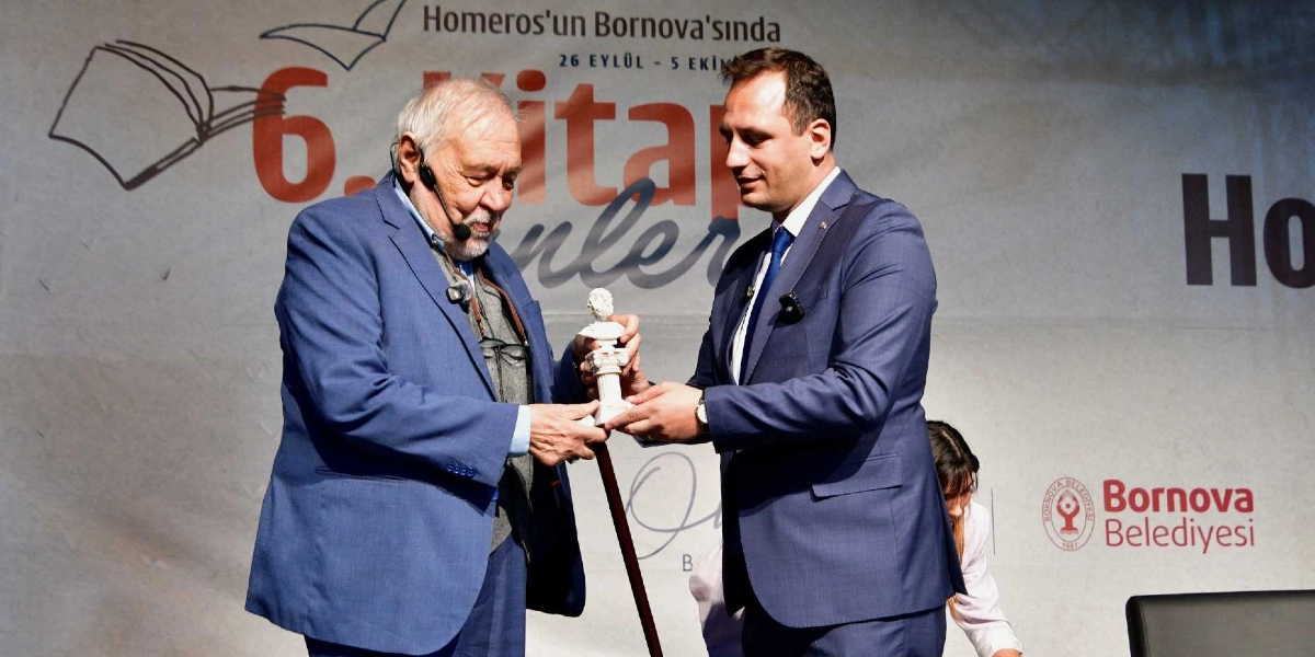 İlber Ortaylı, Bornova Belediyesi tarafından düzenlenen 6. Kitap Günleri etkinliğinde, kendisine takdim edilen Homeros büstünü kabul ederken görülüyor. Ortaylı mavi bir ceket ve gri yelek giymiş, yakasında mikrofon takılı şekilde duruyor. Karşısında lacivert takım elbiseli ve kravatlı bir yetkili, büstü ona uzatıyor. Arka planda 