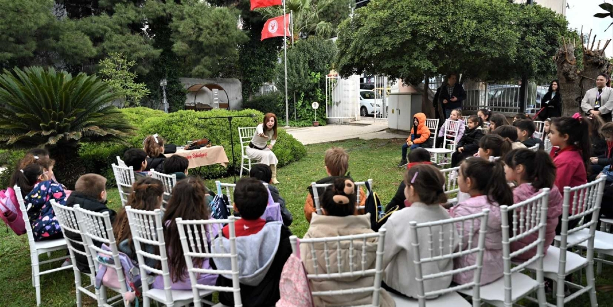Açık havada, yeşil bir bahçede beyaz sandalyelere oturmuş bir grup ilkokul çağındaki çocuk, karşılarında duran ve onlara hitap eden bir kadını dinliyor. Kadın, üzerinde Bornova Belediyesi yazılı bir örtü bulunan küçük bir masanın yanındaki sandalyede oturuyor. Arka planda ağaçlar, çitler ve direklerde asılı Türk bayrakları ile Bornova Belediyesi bayrağı görülüyor.