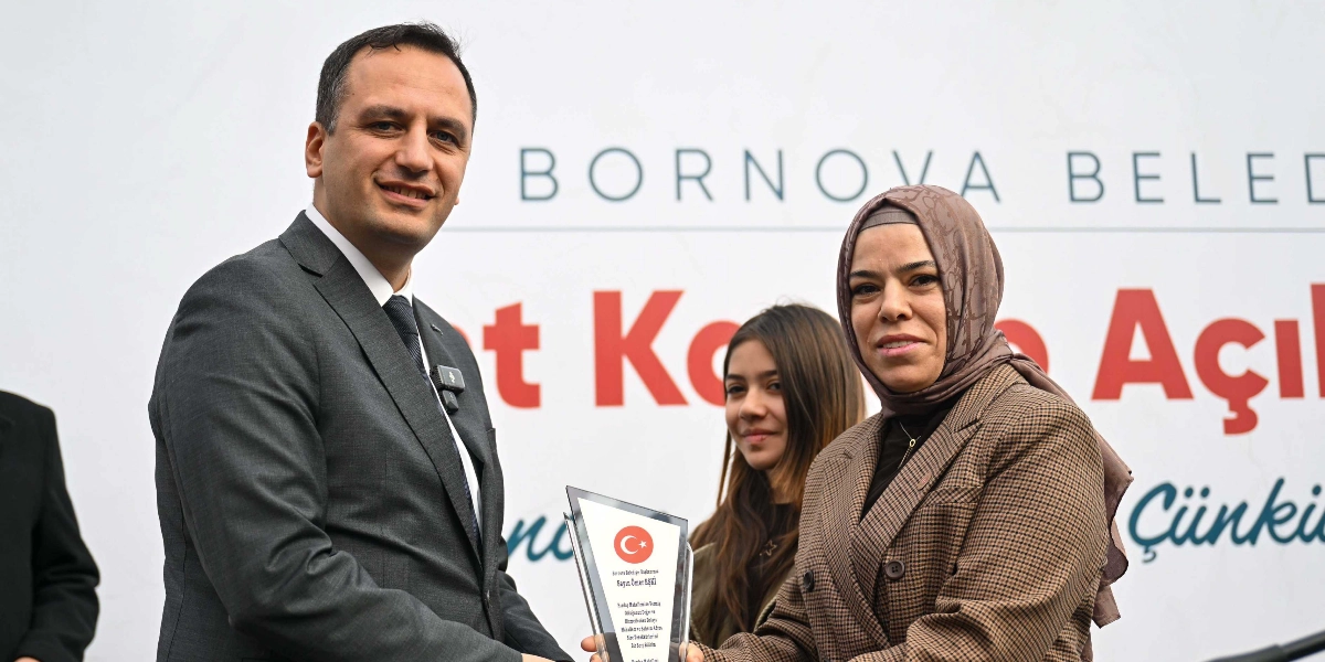 Bornova Belediye Başkanı Ömer Eşki, üzerinde Türk bayrağı ve teşekkür metni bulunan bir plaketi, başörtülü bir kadına takdim ediyor. Arka planda 