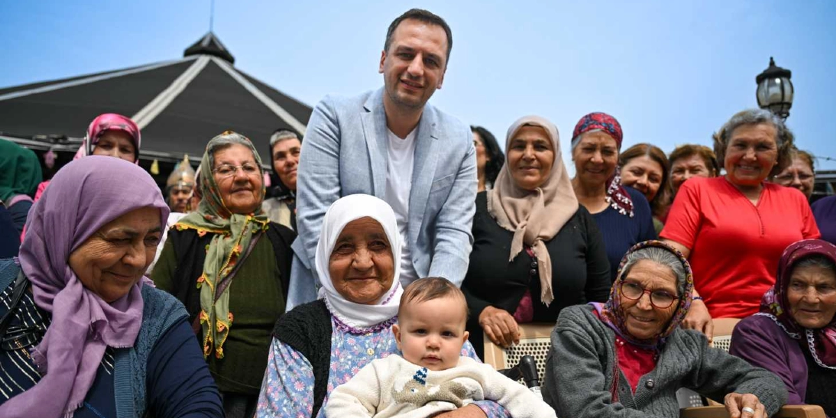 Açık renkli ceket giymiş bir erkek, Anneler Günü kutlaması kapsamında aralarında başörtülü kadınların da bulunduğu kalabalık bir grupla birlikte gülümsüyor. Grubun ön tarafında, beyaz başörtülü yaşlı bir kadının kucağında küçük bir bebek oturuyor. Arka planda ise dış mekan bir etkinlik alanı ve gökyüzü görülüyor.