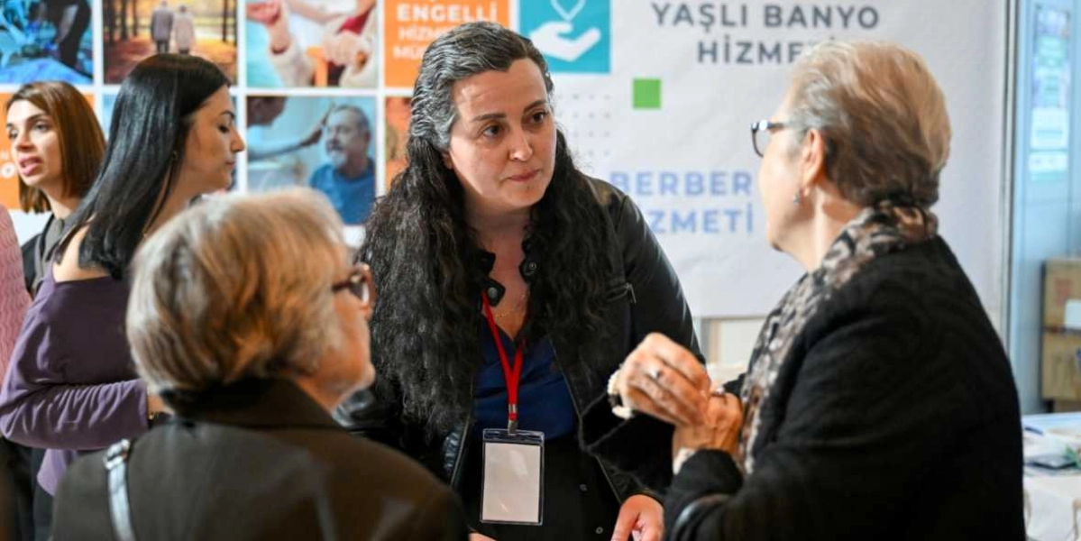 Koyu renkli deri ceket giymiş, boynunda yaka kartı bulunan uzun saçlı bir kadın, yaşlı bir kadınla sohbet ediyor. Arka planda, üzerinde 