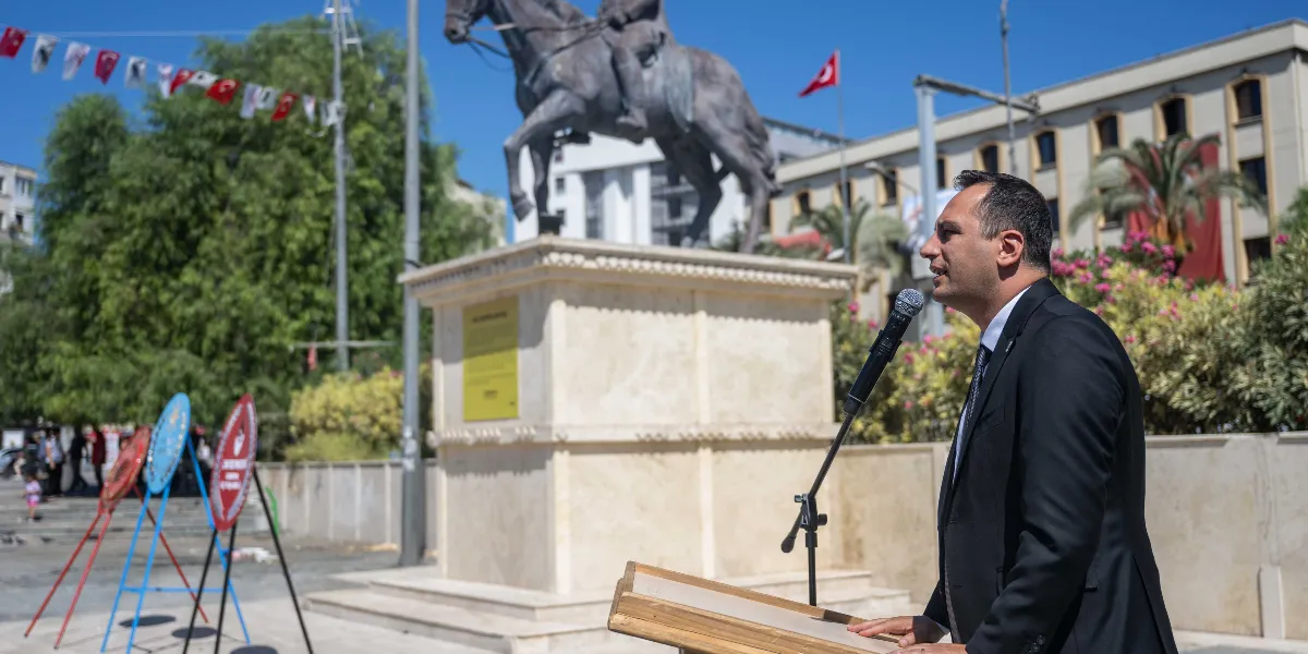 Açık havada, arkasında Atatürk’ün at üzerindeki heykelinin bulunduğu bir meydanda, kürsü başında konuşma yapan takım elbiseli bir erkek. Arka planda Türk bayrakları ve tören çelenkleri yer alıyor.
