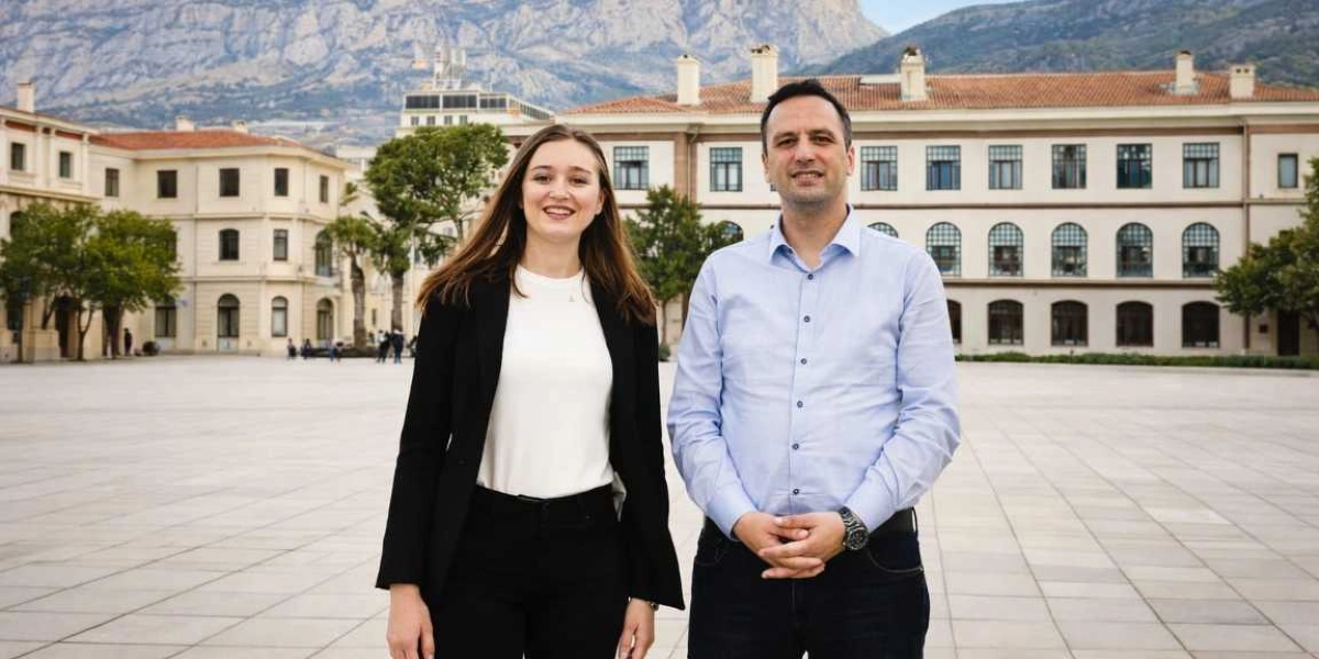 Gülşah Durbay ve yanında duran bir erkek, arkalarında tarihi bir bina ve dağ manzarası bulunan geniş, taş döşeli bir meydanda yan yana durarak gülümsüyor.