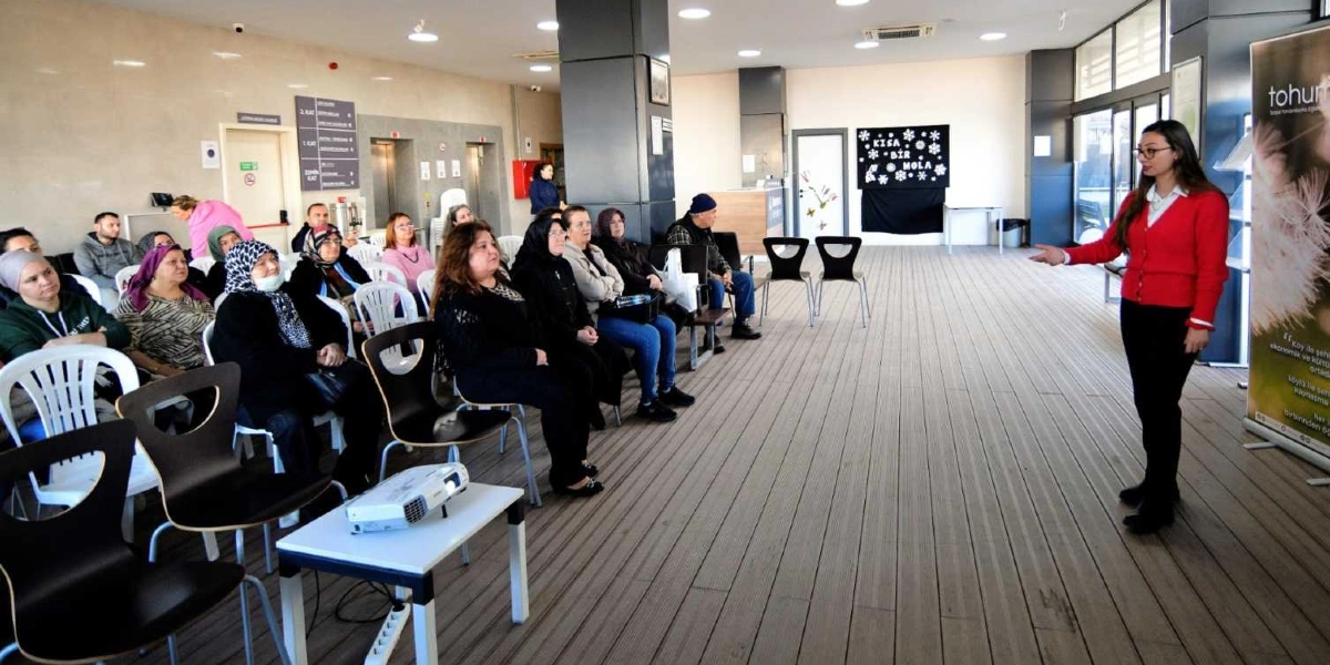 Modern bir binanın geniş salonunda, kırmızı hırkalı bir kadın ayakta durarak katılımcılara sunum yapıyor. Karşısında beyaz sandalyelerde oturan, çoğunluğu kadınlardan oluşan bir grup dinleyici yer alıyor. Salonun önünde bir projeksiyon cihazı bulunurken, arka duvarda üzerinde 