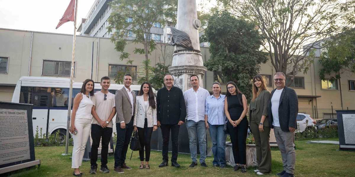 Bornova'da düzenlenen 6. Kitap Günleri kapsamında, bir anıtın önünde yan yana duran dokuz kişilik bir grup. Arka planda beyaz bir minibüs, ağaçlar ve binalar yer alıyor. Grubun her iki yanında üzerinde metinler bulunan bilgilendirme panoları bulunuyor.