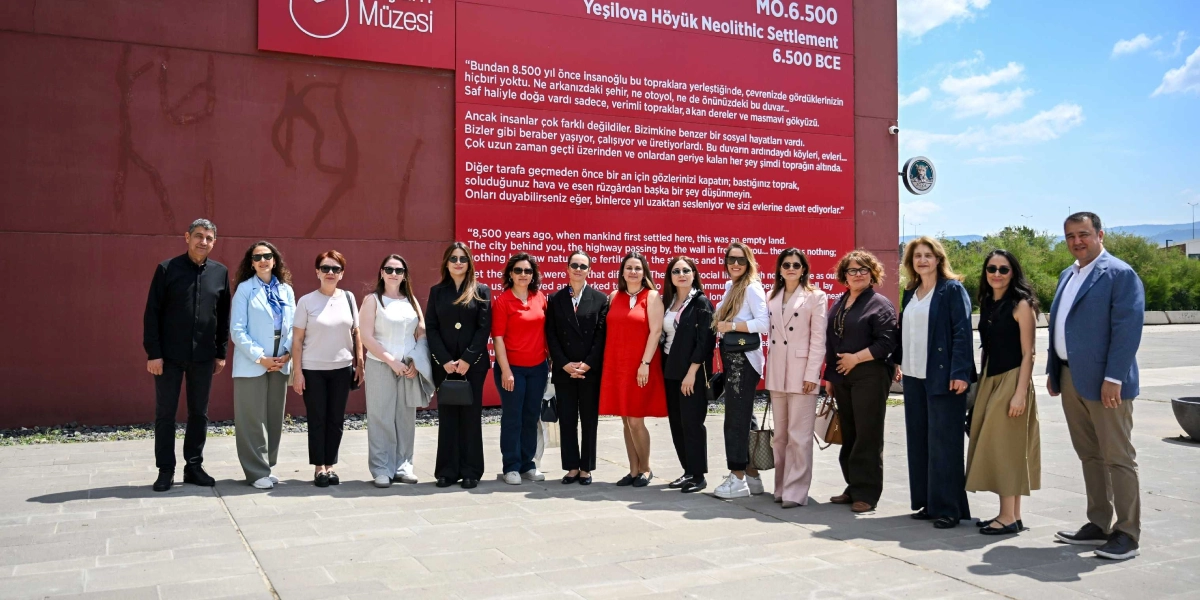 Bornova Yeşilova Höyüğü Ziyaretçi Merkezi'nin kırmızı renkli dış duvarı önünde yan yana duran, aralarında belediye başkan eşlerinin de bulunduğu kalabalık bir grup. Arka plandaki duvarda, Yeşilova Höyüğü Neolitik Yerleşimi hakkında Türkçe ve İngilizce bilgilendirme metinleri yer alıyor. Grup üyeleri günlük ve şık kıyafetler içinde, güneşli bir havada objektife poz veriyor.