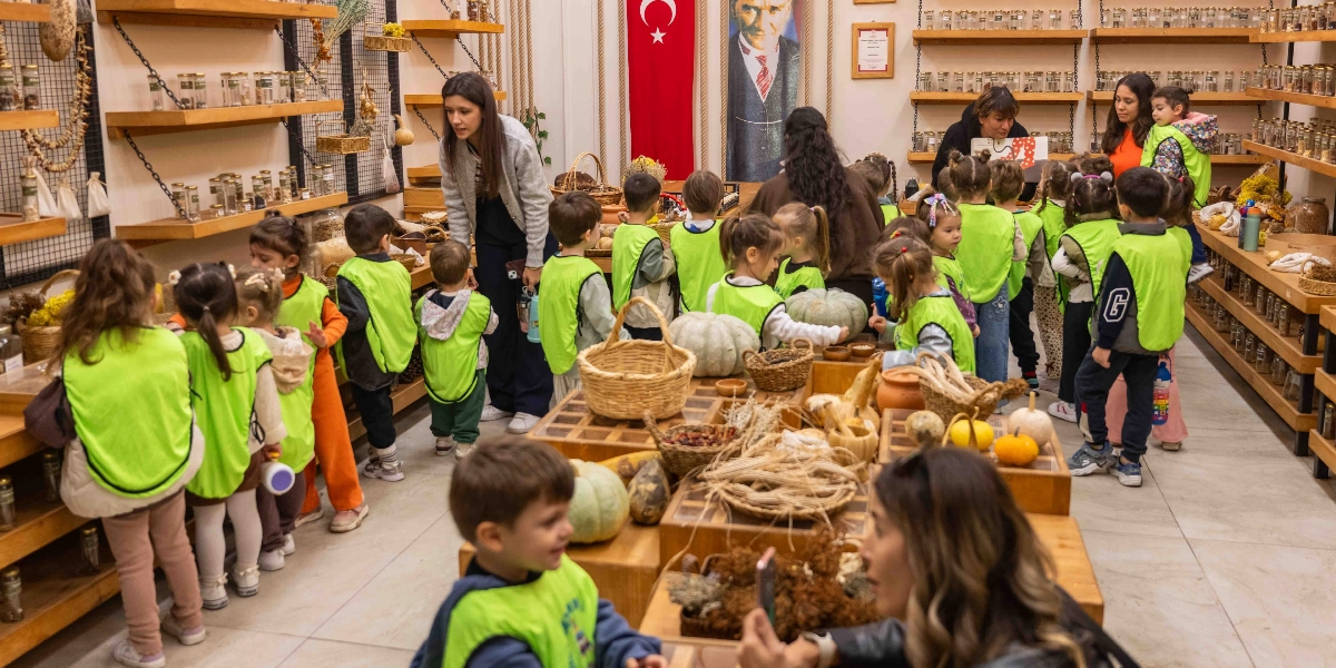 Neon yeşil yelekler giymiş bir grup anaokulu öğrencisi, raflarında çok sayıda kavanoz ve doğal ürün bulunan bir dükkanda öğretmenleri eşliğinde geziyor. Çocuklar ahşap tezgâhların üzerindeki kabakları ve bitkisel ürünleri incelerken, arka planda duvarda asılı Türk bayrağı ve Mustafa Kemal Atatürk posteri dikkat çekiyor. Öğretmenler çocuklara rehberlik ederken, bazı çocuklar merakla etrafı gözlemliyor.