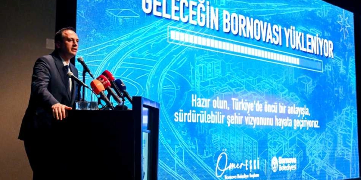 Koyu renk takım elbiseli bir konuşmacı, önündeki kürsüde bulunan mikrofonlara hitap ediyor. Konuşmacının arkasındaki dev ekranda mavi tonlarda bir şehir silüeti arka planı ve üzerinde 