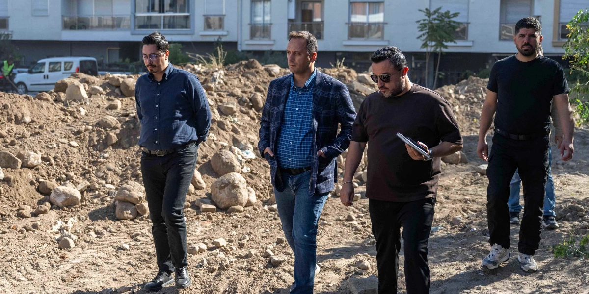 Dört erkek, inşaat halindeki veya düzenleme yapılacak toprak bir arazide yan yana yürürken görülüyor. En soldaki kişi lacivert gömlek ve siyah pantolon giyiyor. Yanındaki kişi kareli bir ceket ve mavi kot pantolonla, onun yanındaki kişi ise kahverengi tişört ve siyah pantolonla yürüyor; elinde küçük bir not defteri tutuyor. En sağda ise siyah tişört ve siyah pantolon giyen bir kişi yer alıyor. Arka planda ise çok katlı apartmanlar ve beyaz bir araç seçiliyor.