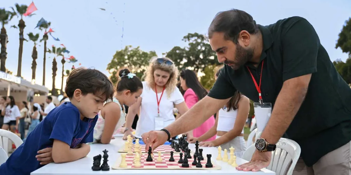 İzmir Enternasyonal Fuarı'ndaki Bornova Belediyesi standında, koyu yeşil tişörtlü bir eğitmen, satranç masasında karşısında oturan küçük bir çocuğa hamle yaparken rehberlik ediyor. Masanın etrafında başka çocuklar ve onları izleyen yetişkinler bulunuyor; arka planda ise ağaçlar ve fuar alanındaki süslemeler yer alıyor.