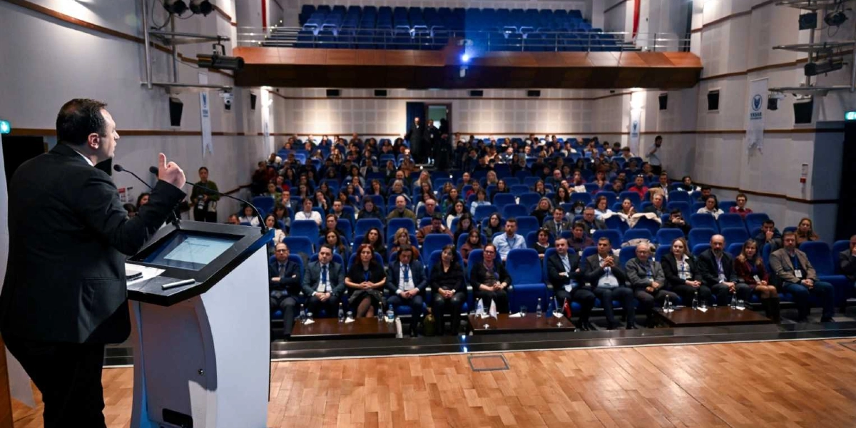 Modern bir konferans salonunda kürsüde konuşma yapan bir erkek ve onu dinleyen kalabalık bir izleyici grubu. Konuşmacı, salonun sol tarafında yer alan kürsüde duruyor ve el hareketleriyle sunumunu gerçekleştiriyor. Salonun geri kalanında, mavi koltuklarda oturan çok sayıda katılımcı dikkatle konuşmacıyı dinliyor. Salonun ön tarafında, izleyicilerin önünde küçük masalar bulunuyor ve duvarlarda Yaşar Üniversitesi'ne ait logolu flamalar asılı.