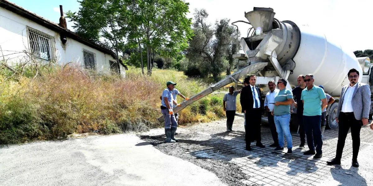 Bornova Belediyesi ekiplerinin kırsal bir alanda beton mikseri kullanarak yol yapım çalışması yürüttüğü sırada, takım elbiseli yetkililer ve çalışanlar devam eden inşaatı inceliyor.