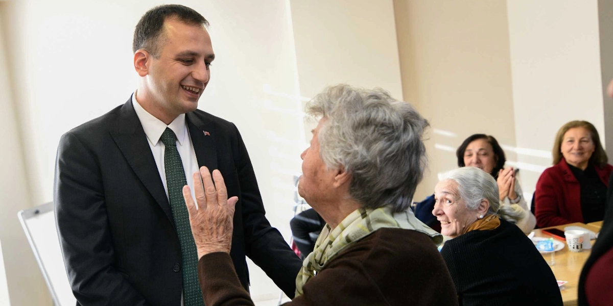 Koyu renk takım elbiseli ve yeşil kravatlı bir erkek, elini havaya kaldırarak selam veren yaşlı bir kadınla gülümseyerek sohbet ediyor. Arka planda ise bir masada oturup gülümseyerek onları izleyen diğer yaşlı kadınlar bulunuyor.