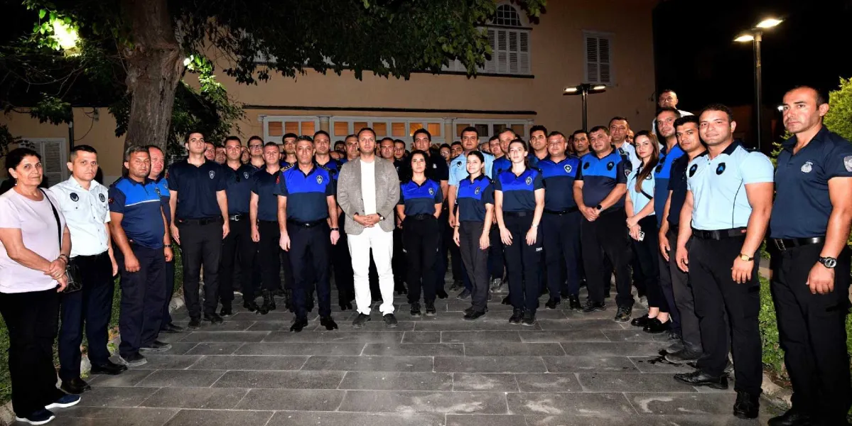 Gece vakti dış mekanda, resmi üniformalı zabıta personeli ve sivil kıyafetli bir erkekle birlikte yan yana duran kalabalık bir grup. Grubun merkezinde bej renkli ceket ve beyaz pantolon giyen bir erkek yer alırken, çevresindeki zabıta personeli mavi ve lacivert renkli üniformalarıyla sıralanmış şekilde kameraya bakıyor. Grubun en solunda ise beyaz tişörtlü bir kadın bulunuyor. Arka planda ağaçlar ve aydınlatılmış bir bina yapısı görülüyor.