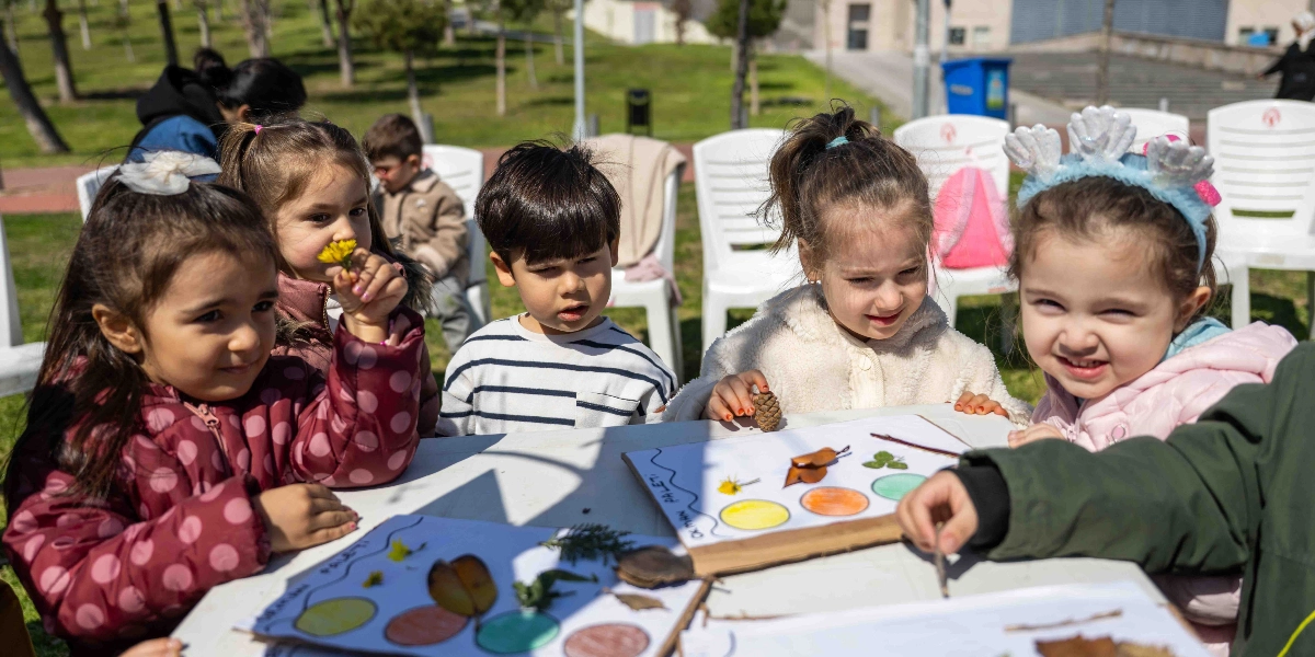 Açık havada, beyaz masaların etrafında toplanmış beş çocuk, doğadan topladıkları yaprak, çiçek ve kozalak gibi malzemeleri kullanarak kağıt üzerindeki renkli daireleri süslüyor. Çocuklardan biri elindeki sarı çiçeği koklarken, diğeri başında deniz kabuğu şeklinde bir taçla gülümsüyor. Önlerindeki kağıtlarda 