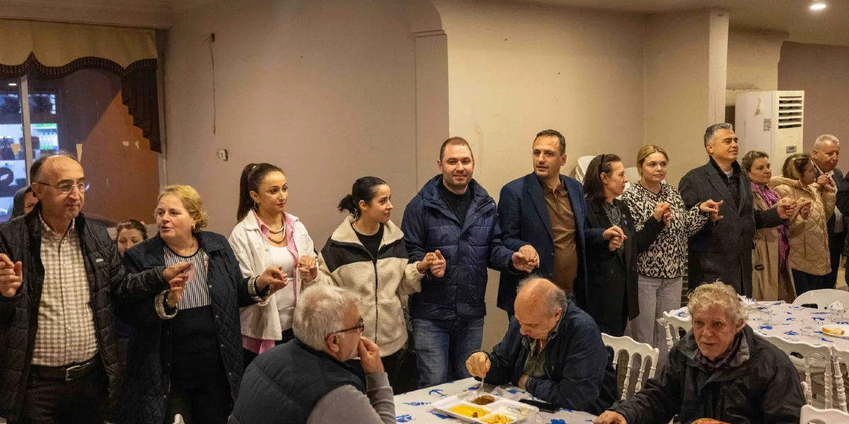 Bornova Belediye Başkanı Ömer Eşki ve bir grup vatandaş, ellerini birbirine kenetleyerek dua etmek için yan yana duruyor. İftar yemeği ortamında çekilen fotoğrafta, ayakta duran grup üyeleri ciddiyetle dua ederken, masada oturan bazı kişiler yemeklerini yemeye devam ediyor. Arka planda sade bir salon dekorasyonu ve yemek masaları görülüyor.