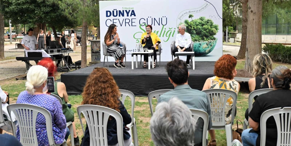 Açık havada, park alanında kurulan bir sahnede düzenlenen 5 Haziran Dünya Çevre Günü Şenliği etkinliğinde, üç kişi sandalyelerde oturarak konuşma yapıyor. Sahnenin arkasındaki büyük panoda 
