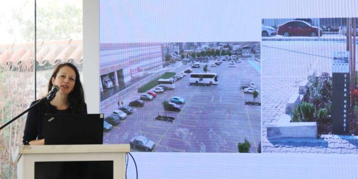 Kürsüde konuşma yapan bir kadın ve arkasındaki dev ekranda yer alan sunum görseli. Ekranda, araçların park halinde olduğu geniş bir otopark alanı ile sağ tarafta 