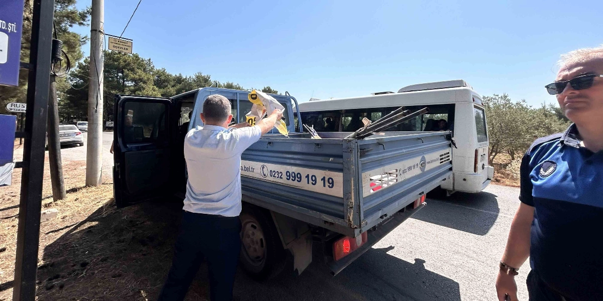 Bornova Belediyesi zabıta ekipleri, yol kenarındaki izinsiz tabelaları ve görsel kirlilik oluşturan materyalleri toplayarak açık kasa bir kamyonete yüklüyor. Sağ tarafta güneş gözlüklü ve üniformalı bir zabıta memuru dururken, diğer bir görevli kamyonetin kasasına metal çubuklar ve tabela parçaları yerleştiriyor. Arka planda beyaz bir minibüs ve ağaçlık bir alan görülüyor.