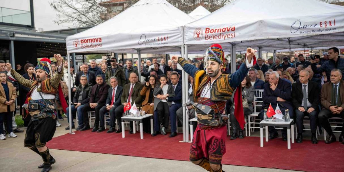 Geleneksel kıyafetler giymiş iki zeybek dansçısı, Bornova Belediyesi logolu beyaz çadırların önünde sergilenen bir etkinlikte performans sergiliyor. Dansçılar, işlemeli yelekleri, başlıkları ve kırmızı şalvarlarıyla kollarını havaya kaldırarak dans ederken, arkalarında protokol üyeleri ve vatandaşlardan oluşan kalabalık bir grup onları izliyor.