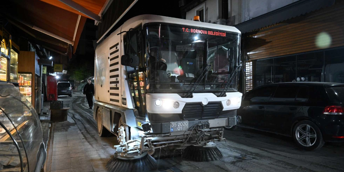 Gece vakti Bornova sokaklarında temizlik yapan T.C. Bornova Belediyesi'ne ait beyaz renkli bir yol süpürme aracı, önündeki döner fırçalarıyla çalışırken görülüyor. Aracın ön camında 