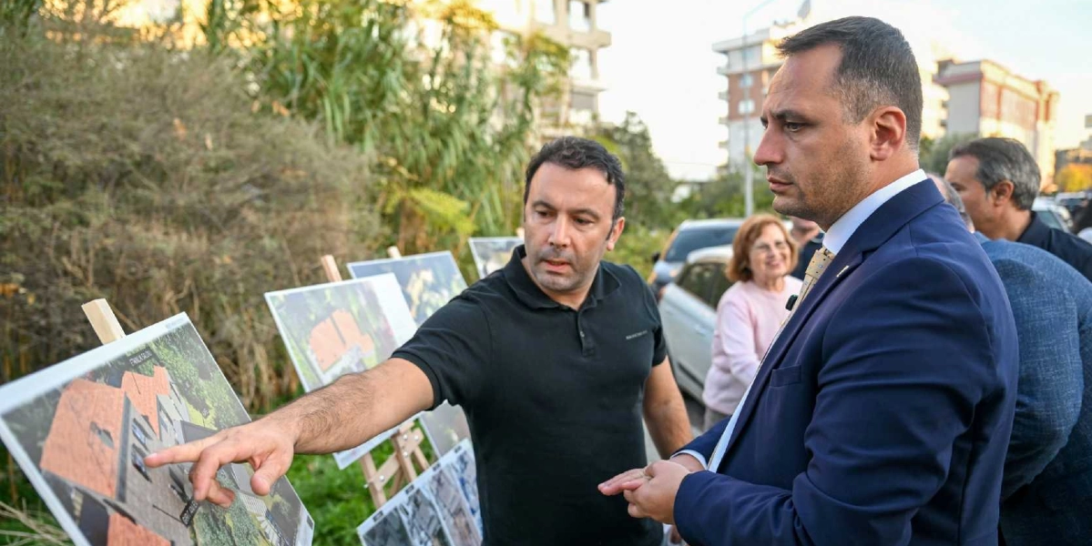 Siyah tişörtlü bir adam, şövale üzerinde sergilenen Girit Anı Evi projesine ait mimari çizimleri lacivert ceketli bir adama eliyle işaret ederek anlatıyor. Arka planda ise bir kadın ve diğer katılımcılar yer alıyor.
