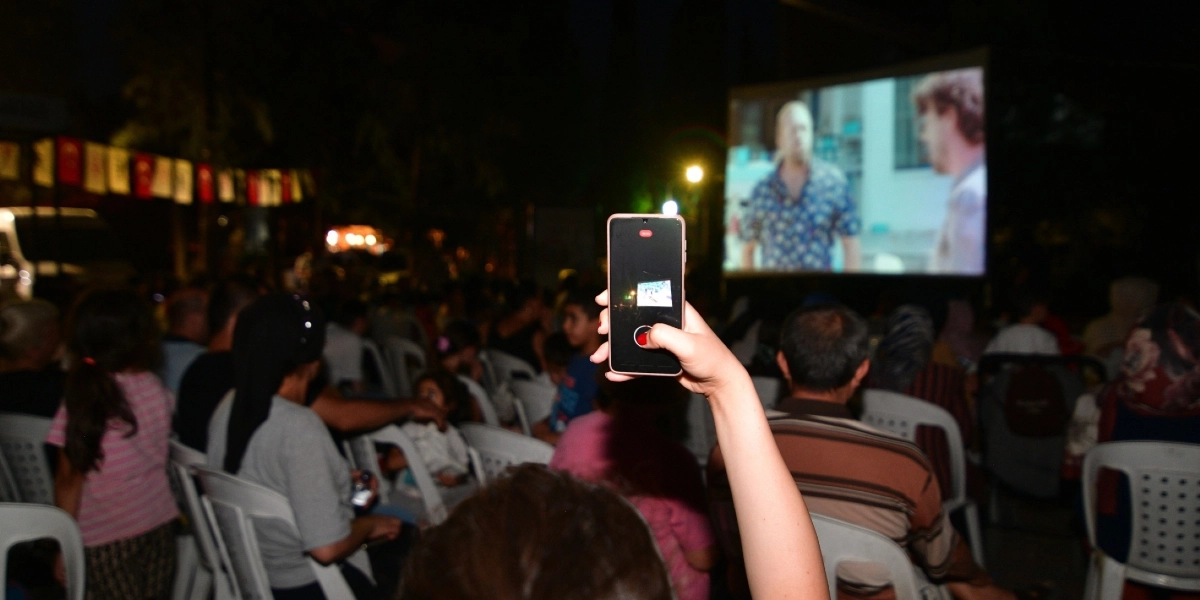 Açık havada kurulan beyaz bir perdeye yansıtılan filmi izleyen kalabalık bir izleyici grubu. Ön planda bir izleyicinin elinde tuttuğu akıllı telefonla perdeyi kaydettiği görülüyor. İzleyiciler beyaz plastik sandalyelerde oturuyor, ortamda akşam saatlerinin karanlığı hakim ve arka planda hafif bir aydınlatma bulunuyor.