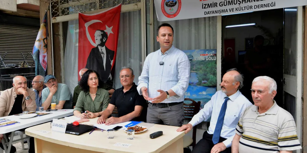 İzmir Gümüşhaneliler Dayanışma ve Kültür Derneği'nde düzenlenen bir toplantıda, açık renkli gömlek giyen bir erkek konuşma yaparken, masanın etrafında oturan katılımcılar onu dinliyor. Arka planda Mustafa Kemal Atatürk'ün portresinin yer aldığı bir Türk bayrağı ve dernek tabelası bulunuyor. Masanın üzerinde 
