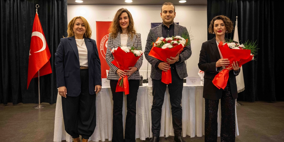 Kapalı bir mekanda, arkalarında Türk bayrağı ve Bornova Belediyesi logosunun bulunduğu bir pankartın önünde yan yana duran dört kişi. Soldan sağa doğru; lacivert ceketli bir kadın, ekose ceketli bir kadın, gri ceketli bir erkek ve siyah ceketli bir kadın yer alıyor. Ortadaki iki kişi ve sağdaki kadın ellerinde kırmızı ambalajlı çiçek buketleri tutuyor. Kişilerin önünde beyaz örtülü bir masa ve üzerinde isimlikler bulunuyor.