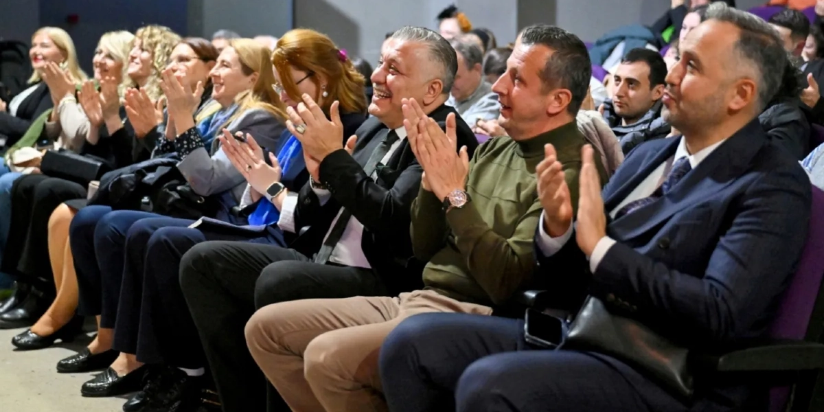 Bir konferans salonunda yan yana oturmuş, gülümseyerek alkışlayan bir grup insan. Ön planda takım elbiseli ve haki renkli kazaklı erkekler, yanlarında ise şık giyimli kadınlar yer alıyor. İzleyiciler, sahnedeki bir etkinliği ilgiyle takip ederek memnuniyetlerini alkışlarla gösteriyor.