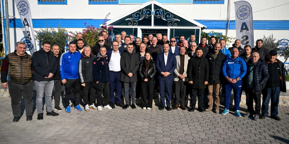 Beyaz ve mavi renkli bir spor tesisi binasının önünde, aralarında Bornova Belediye Başkanı Mustafa İduğ'un da bulunduğu, spor yöneticileri ve temsilcilerden oluşan kalabalık bir grup, toplu halde poz veriyor. Grubun önünde ve arkasında İzmir Amatör Spor Kulüpleri Federasyonu logolu dikey bayraklar yer alıyor. Binanın giriş kapısının üzerinde 