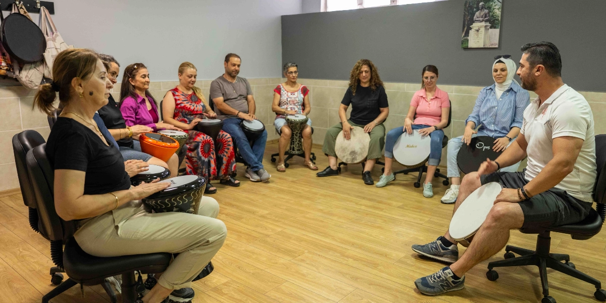 Bir eğitmen eşliğinde çember şeklinde oturmuş, ellerinde darbuka ve benzeri vurmalı çalgılar bulunan bir grup yetişkin, müzik atölyesinde eğitim alıyor.