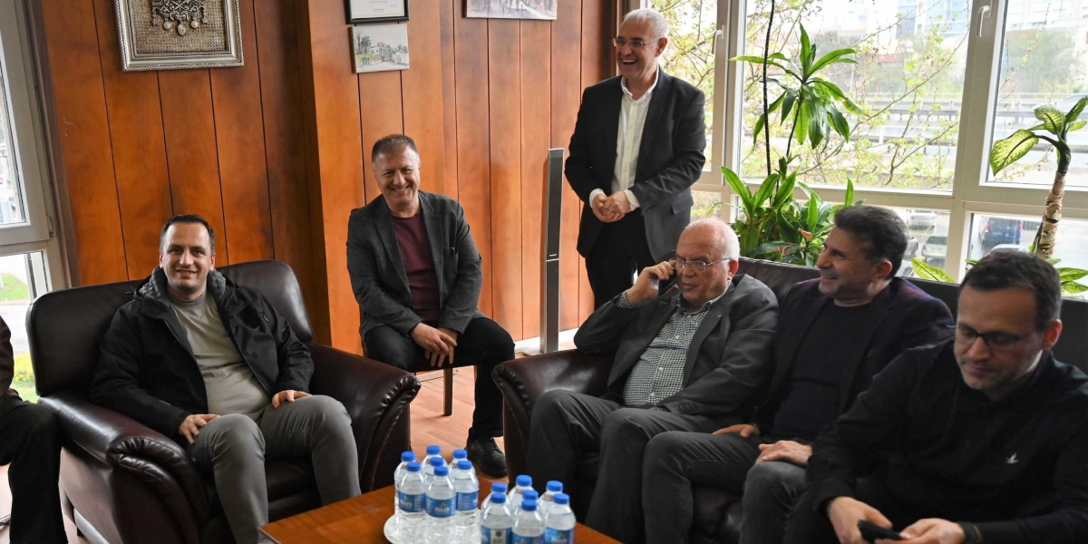 Ahşap kaplamalı bir ofis ortamında, deri koltuklarda oturan ve ayakta duran bir grup erkek samimi bir sohbet içinde. Solda tekli koltukta oturan kişi gülümseyerek bakarken, yanında bordo tişörtlü bir başka kişi daha oturuyor. Sağ taraftaki geniş deri koltukta ise biri telefonla konuşan, diğeri gülümseyen iki kişi yan yana oturuyor; en sağda ise başka bir kişi elindeki telefona bakıyor. Ayakta duran, gri saçlı ve ceketli bir kişi ise gülümseyerek gruba eşlik ediyor. Önlerindeki alçak masanın üzerinde çok sayıda küçük su şişesi bulunuyor.