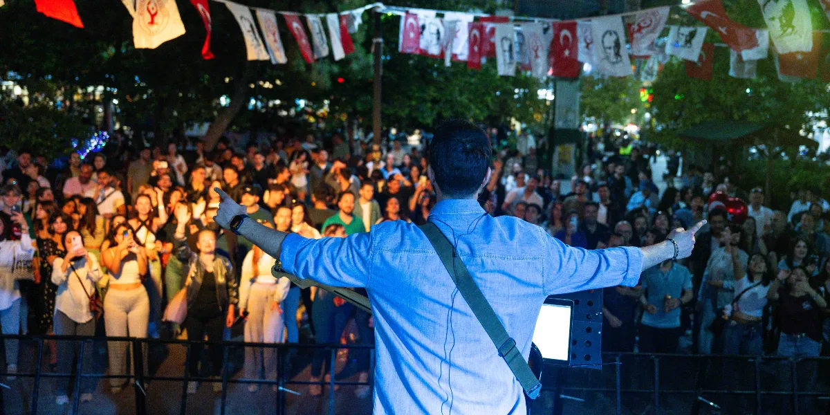 Açık havada düzenlenen bir konserde, sırtı kameraya dönük şekilde elinde gitarıyla sahneden kalabalığa seslenen bir müzisyen ve onu coşkuyla izleyen, ellerini havaya kaldıran gençlerden oluşan büyük bir kitle. Sahne alanının üstü, Türk bayrakları ve Atatürk portrelerinin yer aldığı flamalarla süslenmiş.