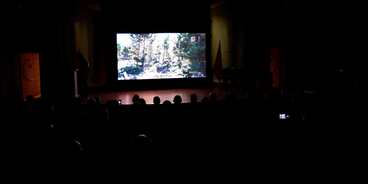 Karanlık bir konferans salonunda, sahnedeki büyük ekranda ormanlık bir alanı gösteren bir belgesel veya tanıtım filmi izleyen izleyiciler.