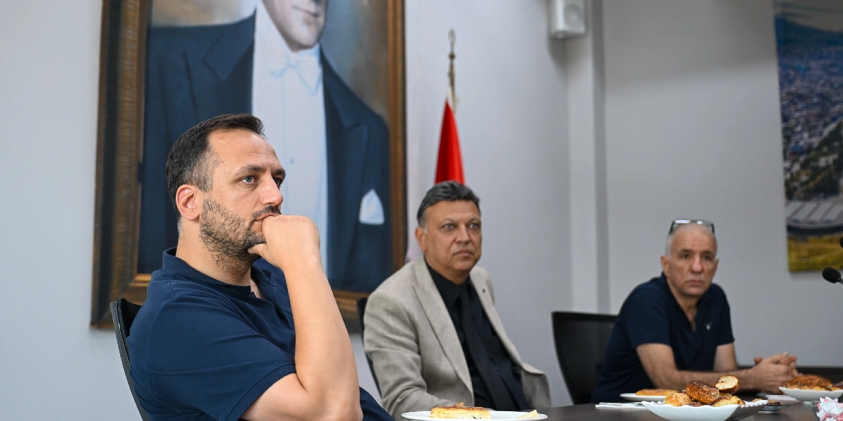 Lacivert tişörtlü bir adam elini çenesine dayamış düşünceli bir şekilde ileriye bakarken, yanında oturan iki kişiyle birlikte bir masada oturuyor. Arka planda duvarda büyük bir Atatürk portresi ve Türk bayrağı yer alıyor. Masanın üzerinde tabaklarda yiyecekler bulunuyor.