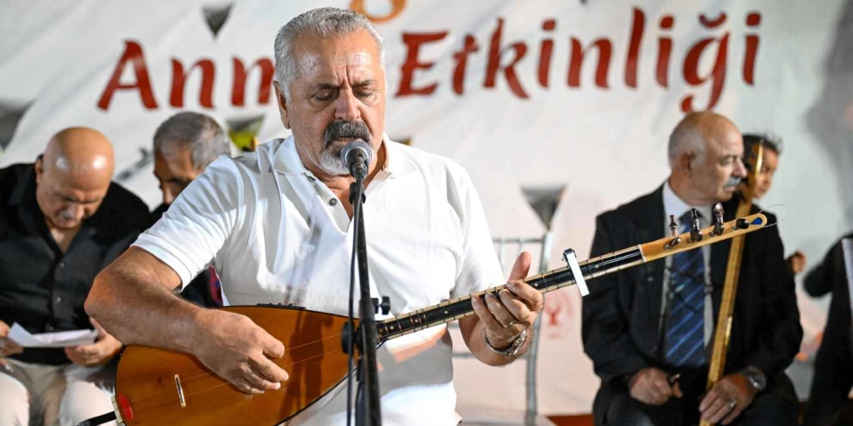 Beyaz polo yaka tişört giymiş, gri saçlı ve bıyıklı bir halk ozanı, elinde bağlamasıyla mikrofon başında şarkı söylüyor. Arka planda üzerinde 