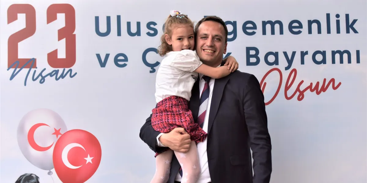 Takım elbiseli bir adam, kucağında beyaz tişörtlü ve kırmızı ekose etekli küçük bir kız çocuğunu tutarak gülümseyerek poz veriyor. Arka planda 