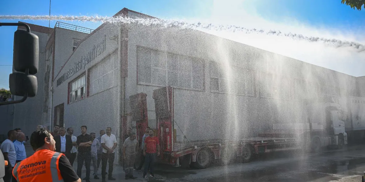 Bornova Belediyesi Şantiyesi binasının önünde, itfaiye ekiplerinin müdahalesiyle havaya sıkılan tazyikli suyun oluşturduğu su perdesi altında, belediye görevlileri ve vatandaşların bulunduğu bir grup insan çalışmaları izliyor.