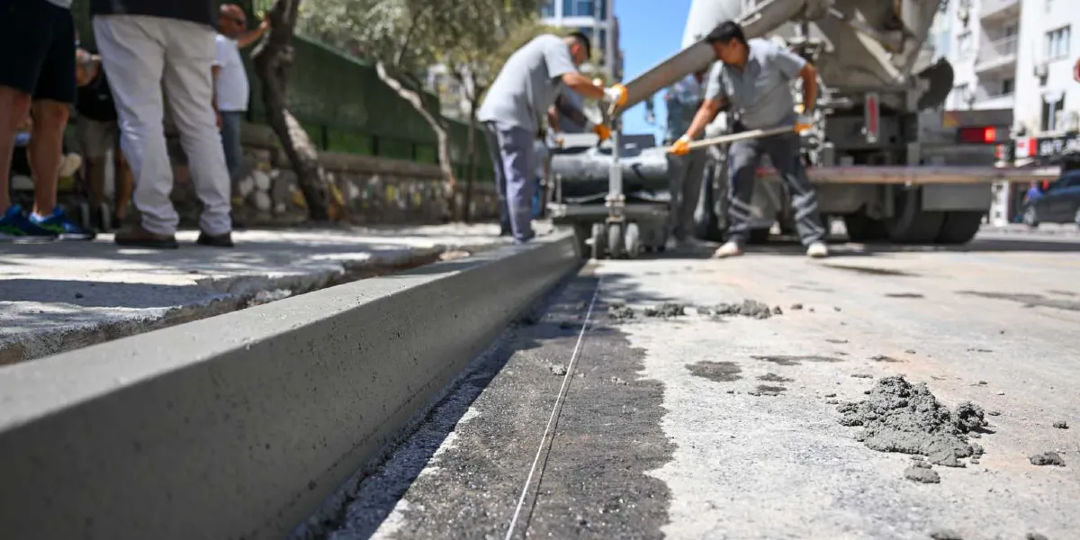 İşçilerin bir beton mikseri yardımıyla yol kenarına yeni bordür taşı döktüğü ve betonlama çalışması yaptığı bir cadde manzarası.
