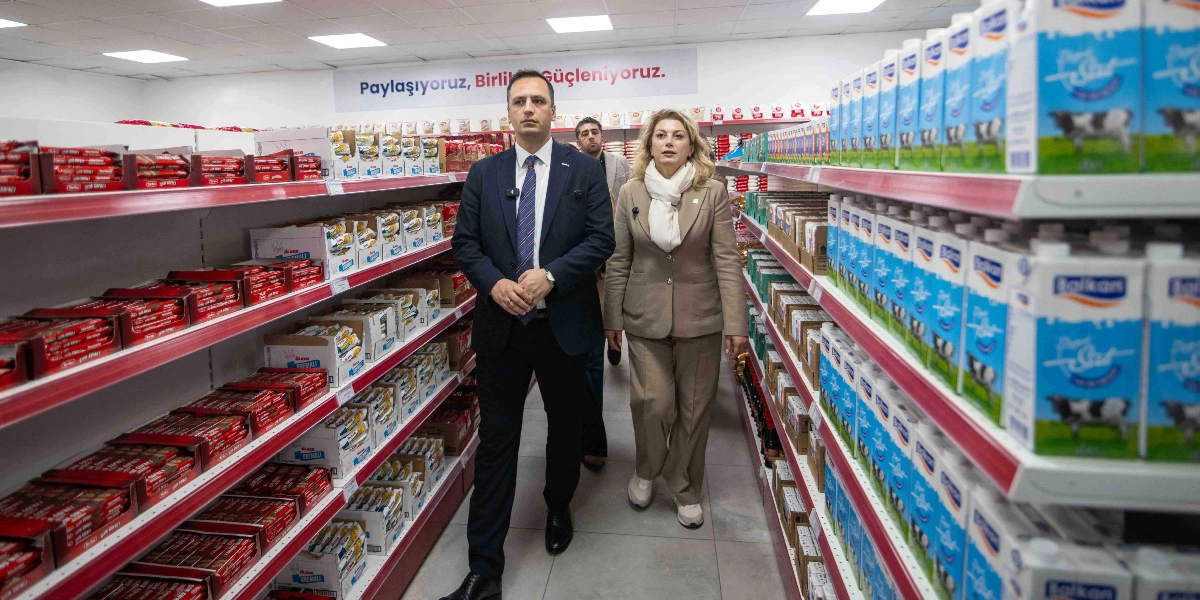 Koyu renk takım elbiseli bir erkek ve bej renkli bir ceket pantolon takımı giyen sarı saçlı bir kadın, raflarında gıda ürünleri dizili bir market reyonunun arasında yan yana yürüyor. Arka planda duvarda 