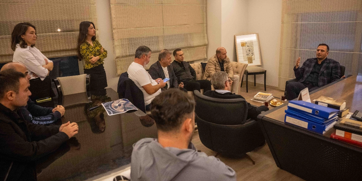 Modern bir ofis ortamında, büyük bir toplantı masasının etrafında toplanmış yaklaşık on kişilik bir grup, bir konu üzerinde görüşme yapıyor. Masanın başında oturan koyu renk ceketli bir kişi konuşurken, diğer katılımcılar onu dinliyor. Masanın üzerinde mavi dosyalar, belgeler ve bir bardak çay bulunuyor. Odanın köşesinde krem rengi bir koltuk takımı yer alıyor ve duvarda çerçeveli bir tablo asılı. Katılımcıların bir kısmı masanın etrafında otururken, bir kısmı ayakta durarak toplantıyı takip ediyor.