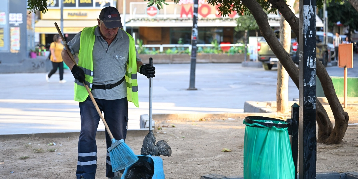 Bornova Belediyesi logolu gri tişört, lacivert pantolon ve sarı reflektörlü yelek giyen bir temizlik görevlisi, elindeki mavi süpürge ve faraşla park alanındaki çöpleri temizliyor. Görevlinin yanında yeşil çöp poşeti takılı bir çöp kovası bulunuyor, arka planda ise ağaçlar ve işletmelerin olduğu bir meydan yer alıyor.