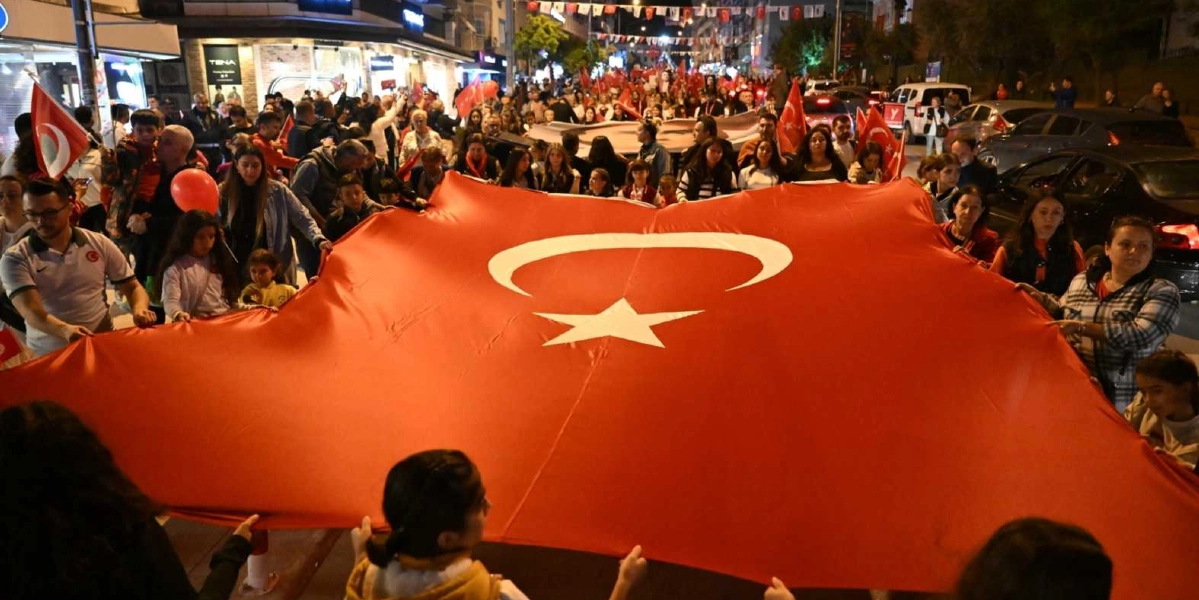 Gece vakti kalabalık bir caddede yürüyen insanlar, ellerinde tuttukları dev bir Türk bayrağını havada taşıyor. Çevredeki katılımcıların birçoğu ellerinde küçük Türk bayrakları tutuyor ve bazıları kırmızı balonlar taşıyor. Sokak ışıklarıyla aydınlanan caddede, coşkulu bir kalabalık 29 Ekim Cumhuriyet Bayramı kutlaması için bir arada yürüyor.